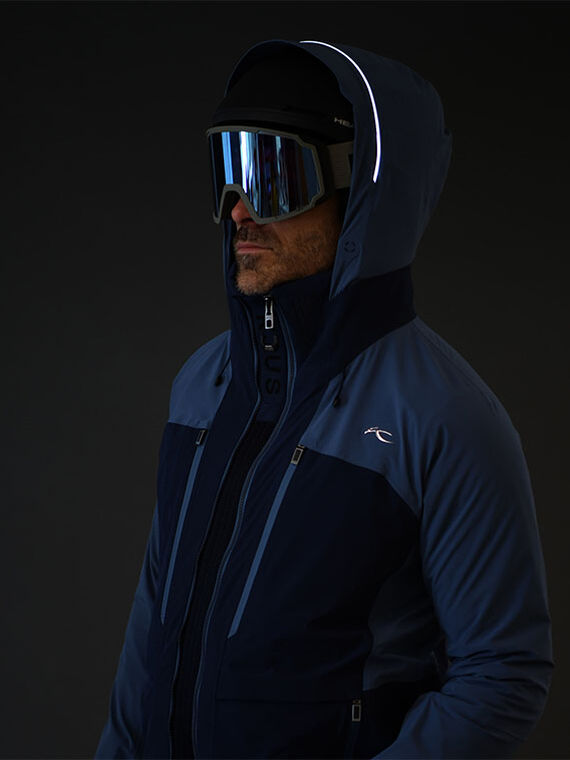 Skier wearing KJUS blue ski jacket with goggles and helmet on a dark studio background