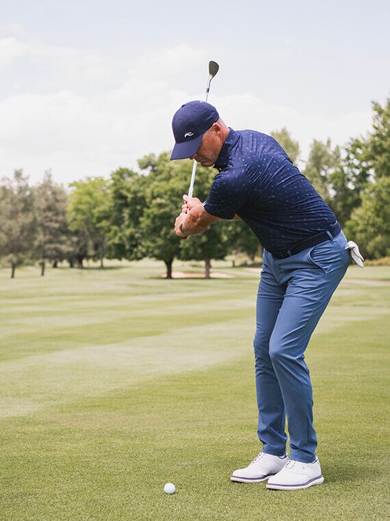 Golfer swinging club on fairway, wearing blue KJUS pants, a navy KJUS polo and cap.