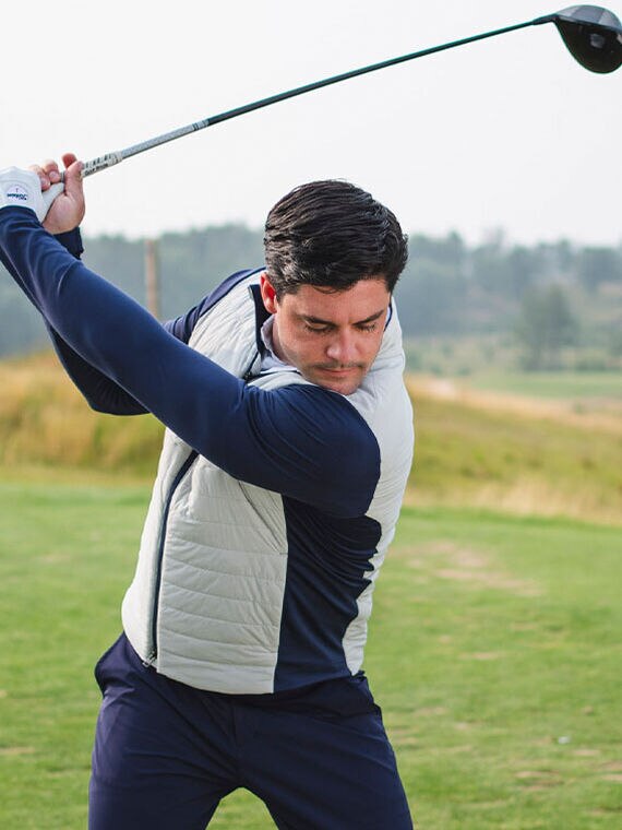 Golfer swinging a club wearing KJUS insulated zip-up jacket and navy pants