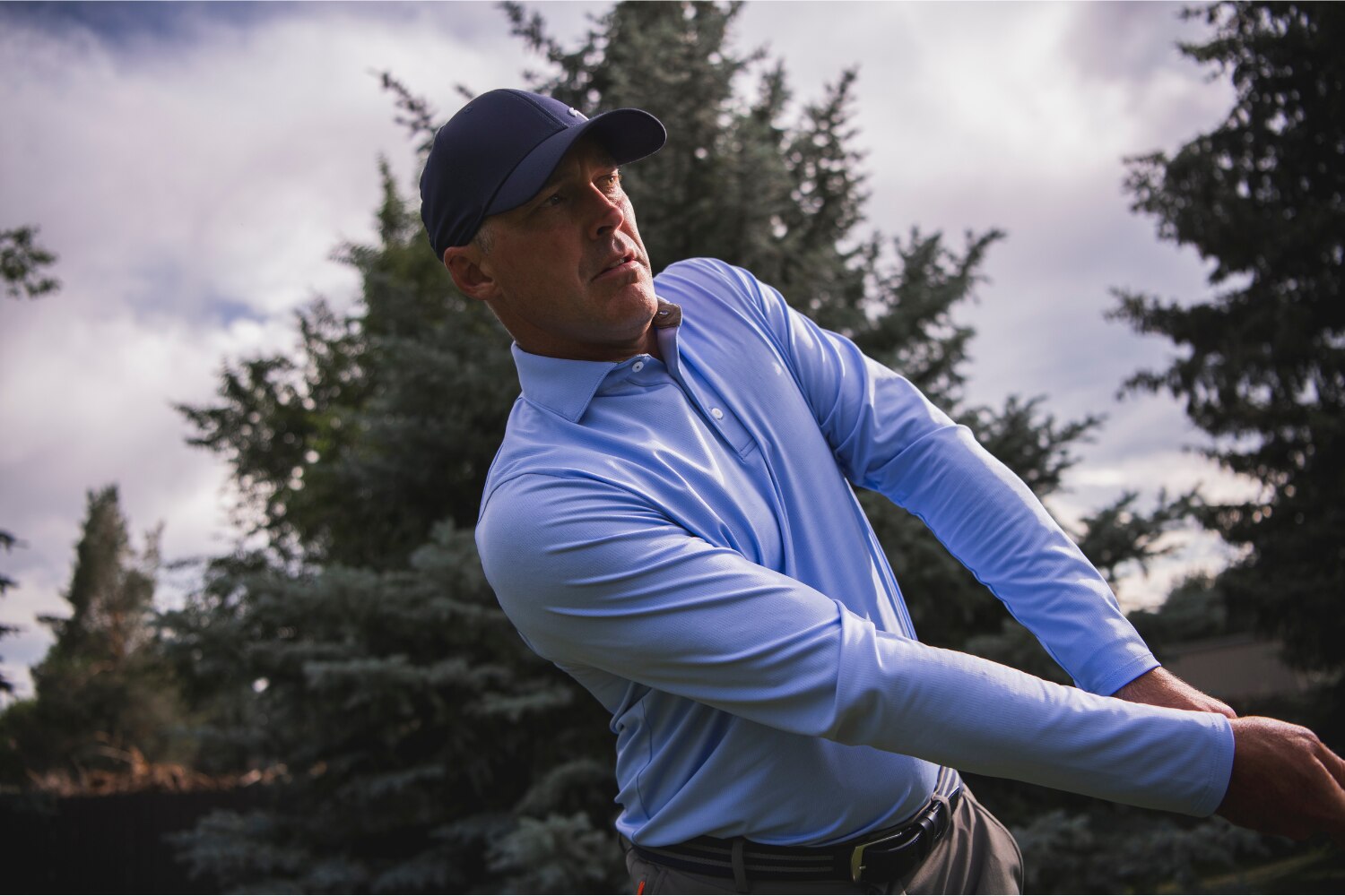 A man wearing a light blue KJUS long-sleeve polo and a navy blue KJUS cap, swinging a golf club.