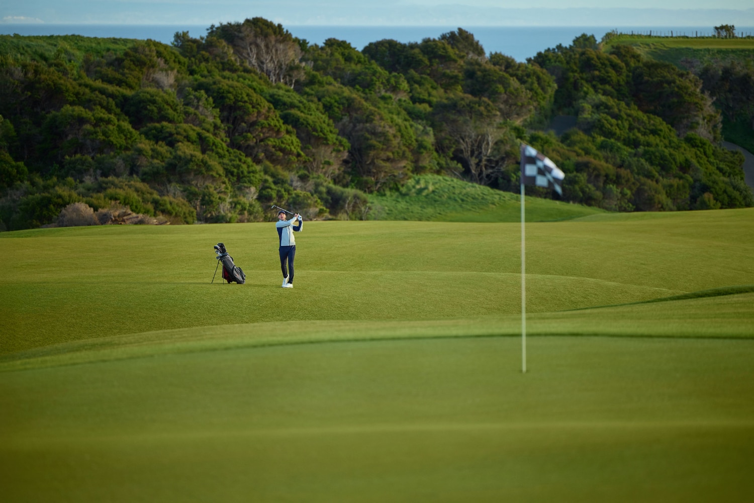A male golf player in full swing on a green, trees in the background.