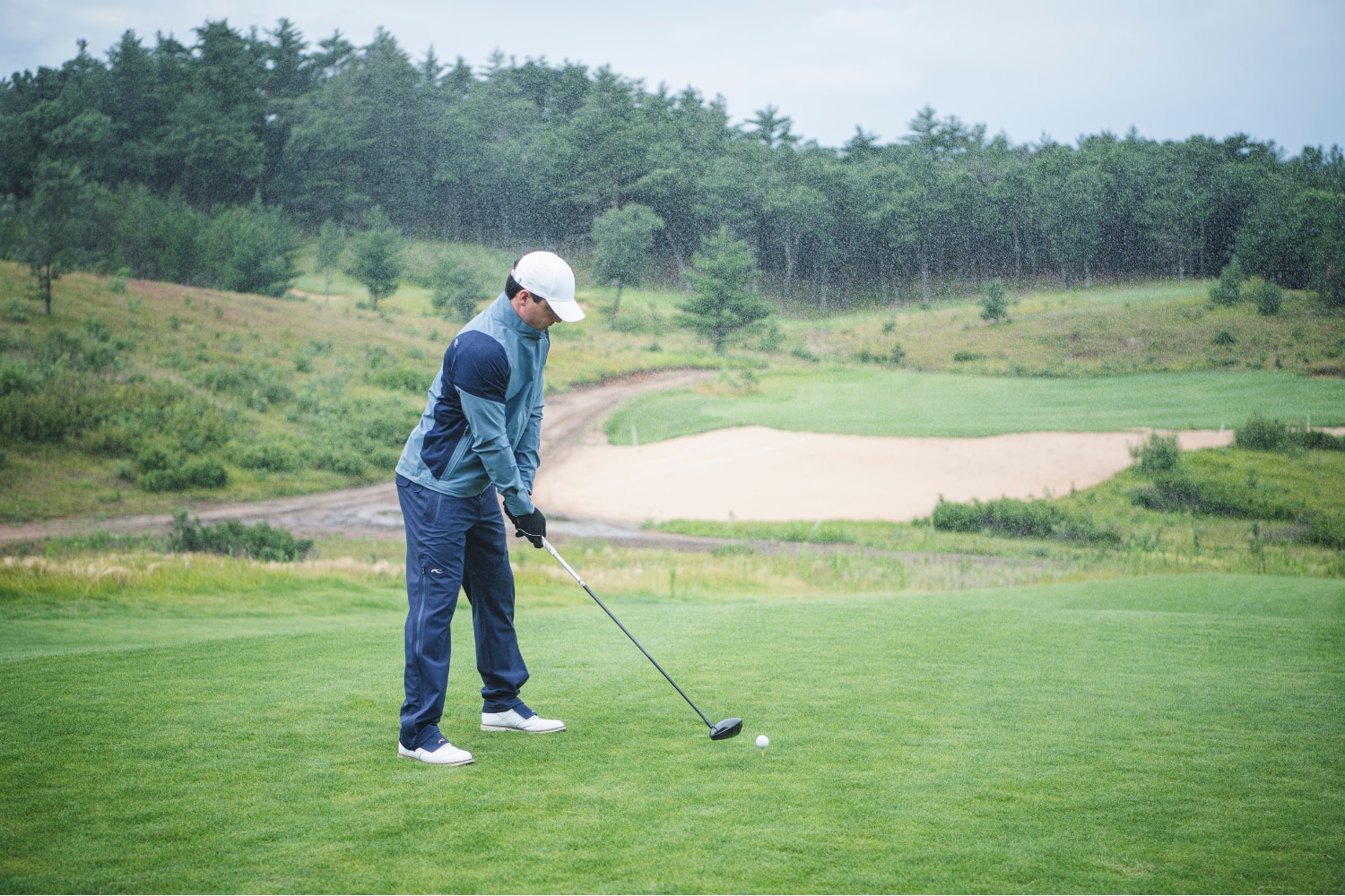 A man wearing a light blue KJUS long-sleeve polo and a navy blue KJUS cap, swinging a golf club.