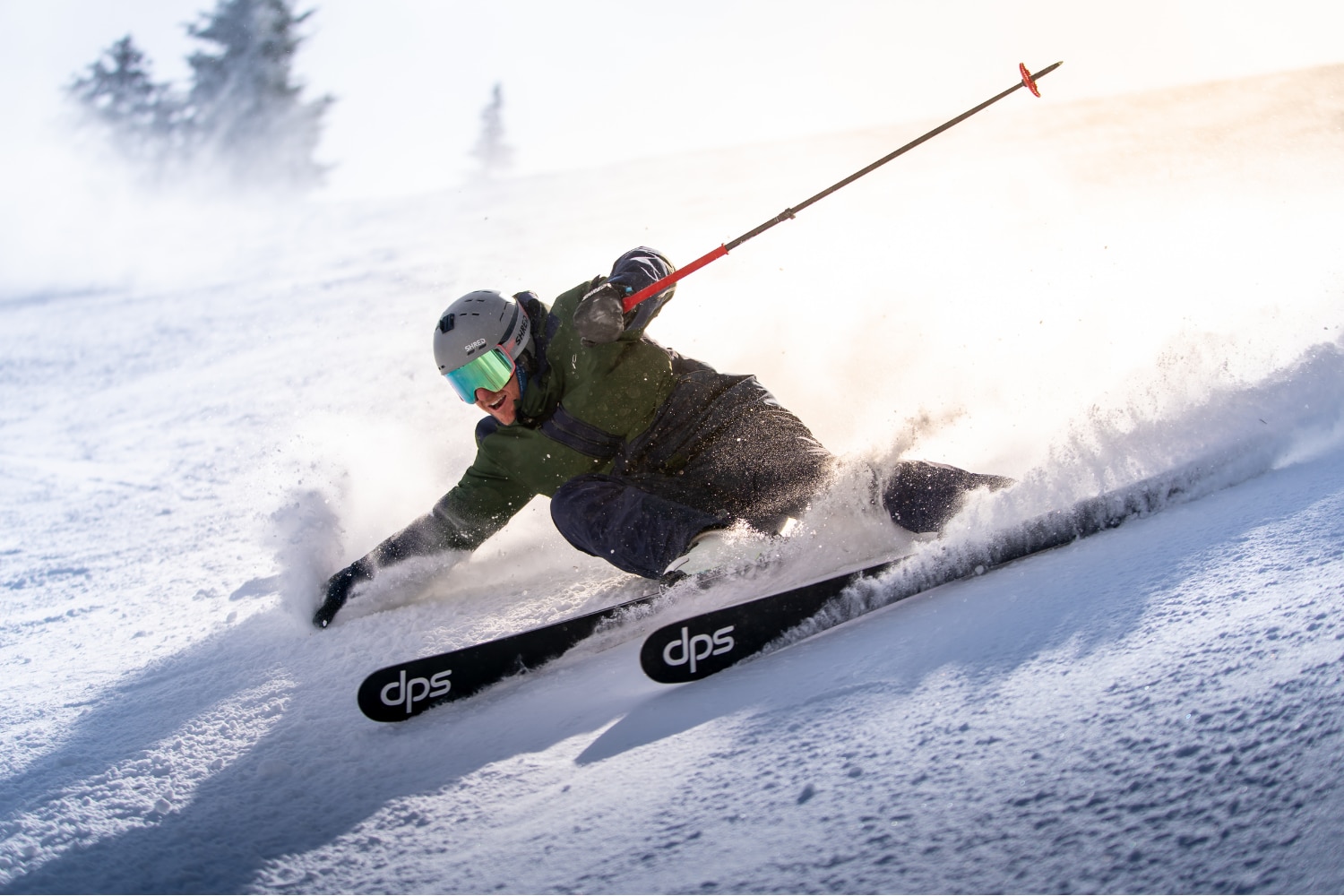 A woman wearing the Women's Twilight Jacket, a helmet and holding her skies over her shoolder in front of a snowy mountain range.