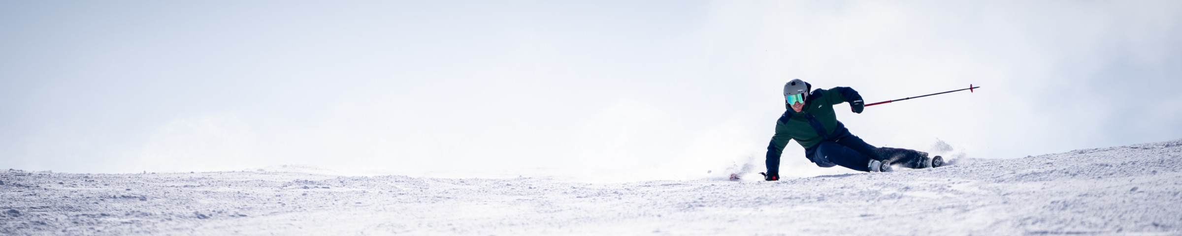 A snowy peak with a skier in a blue KJUS ski outfit, a snowy mountain range in the background.