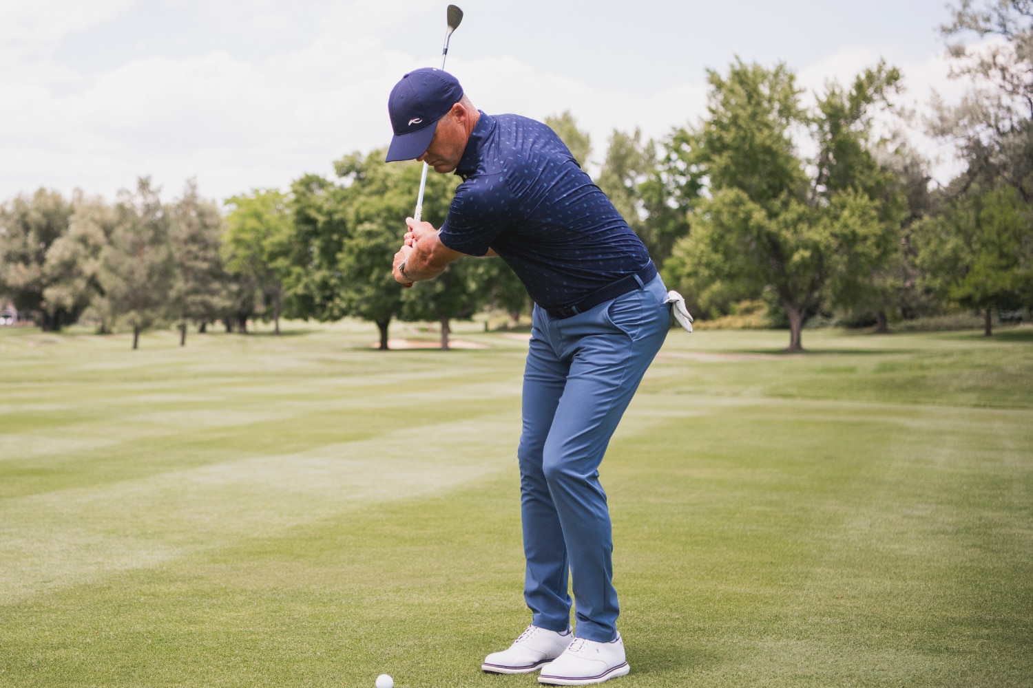 A male golf player wearing a blue KJUS golf outfit in full swing on a green, trees in the background.