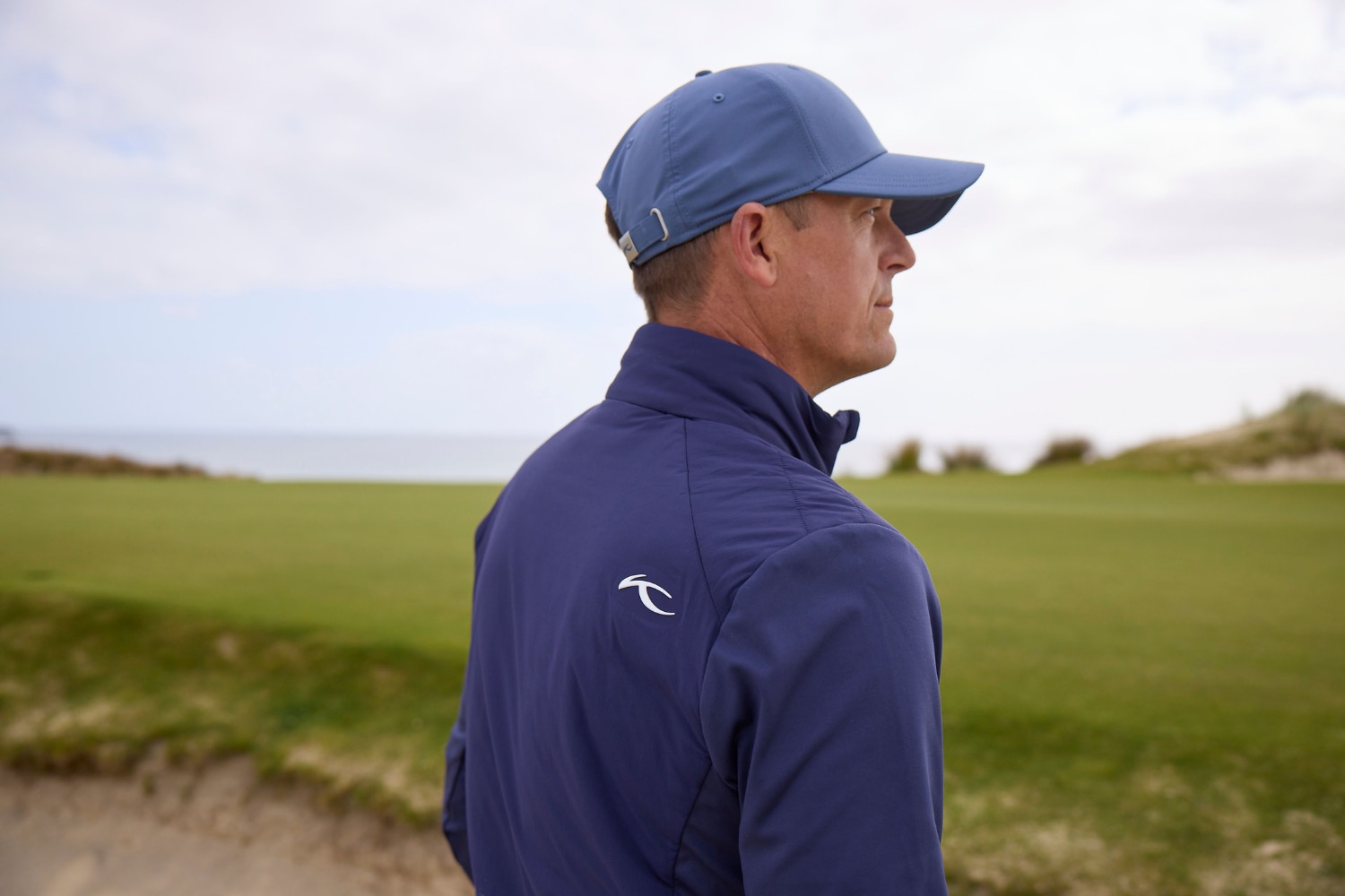 A male golf player with blue KJUS cap and blue Reach jacket looking into the distance.