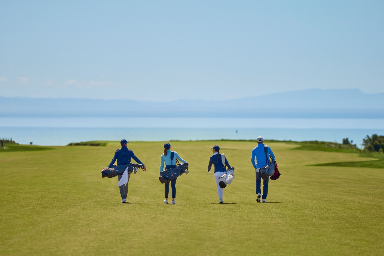 Four golf players walk on a green, the coast and mountains in the background.