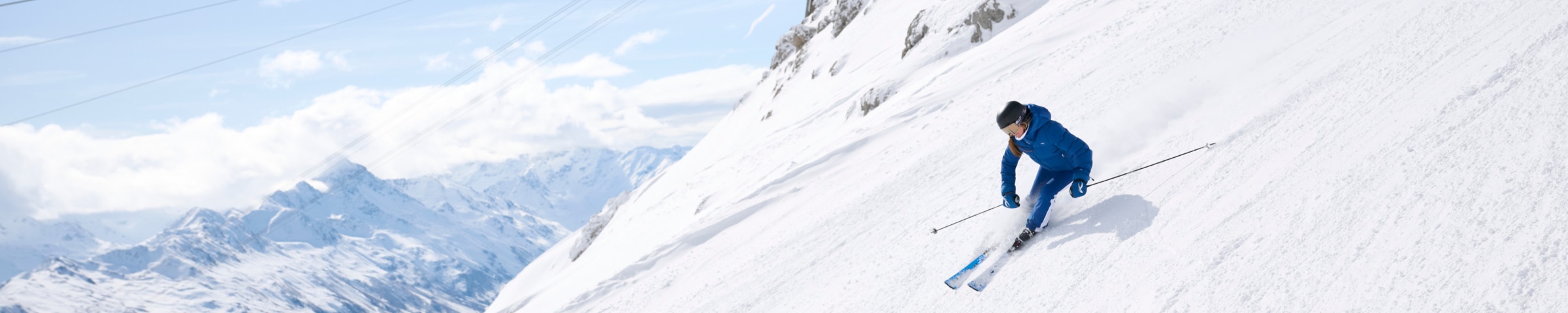 A snowy peak with a female skier in a blue KJUS ski outfit, a snowy mountain range in the background.