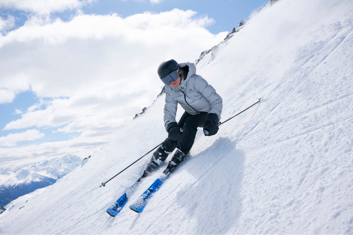 A female skier is racing down a slope, snowy peaks in the background.