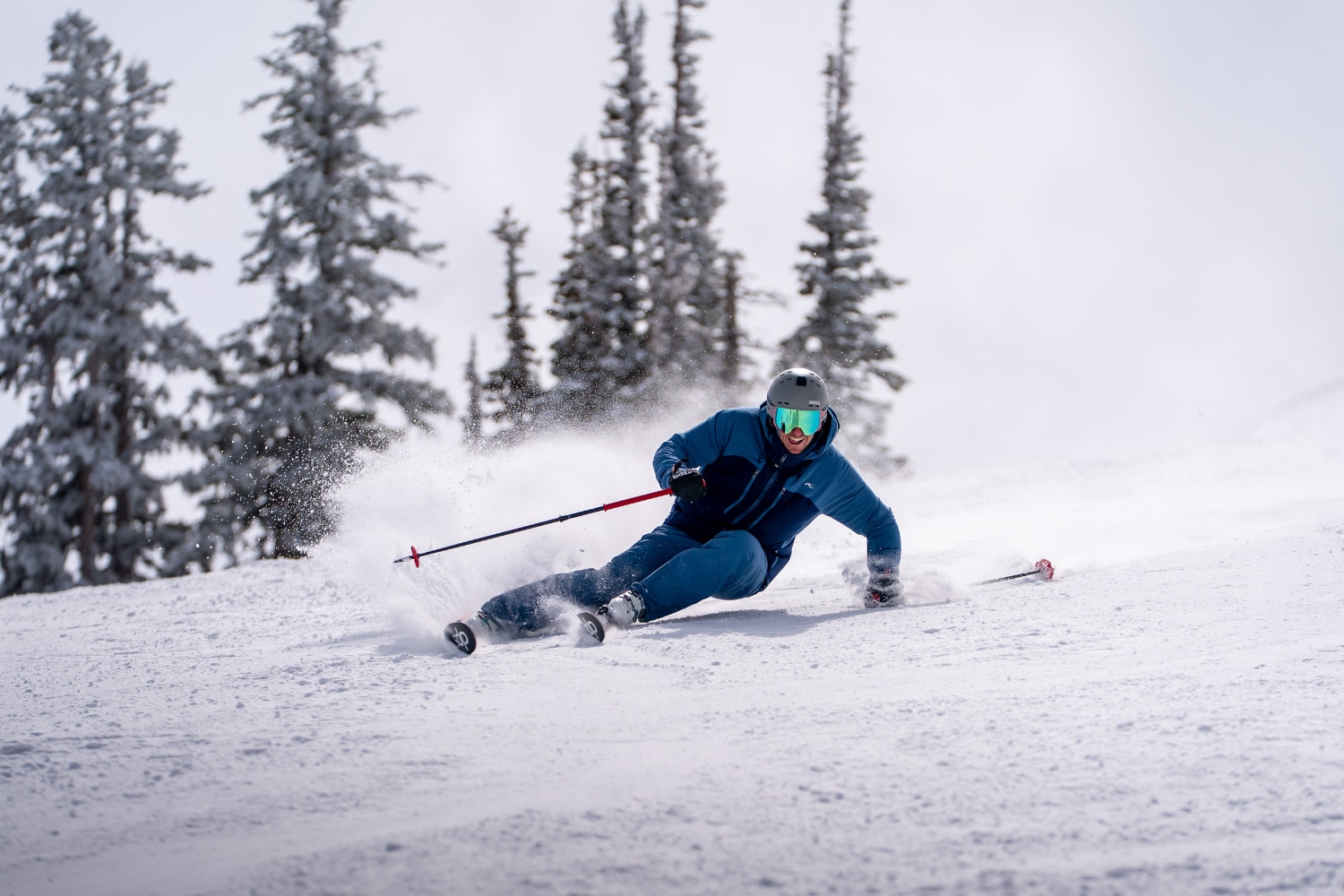 A skier in blue KJUS outfit is carving down a slope, some trees in the backgorund.