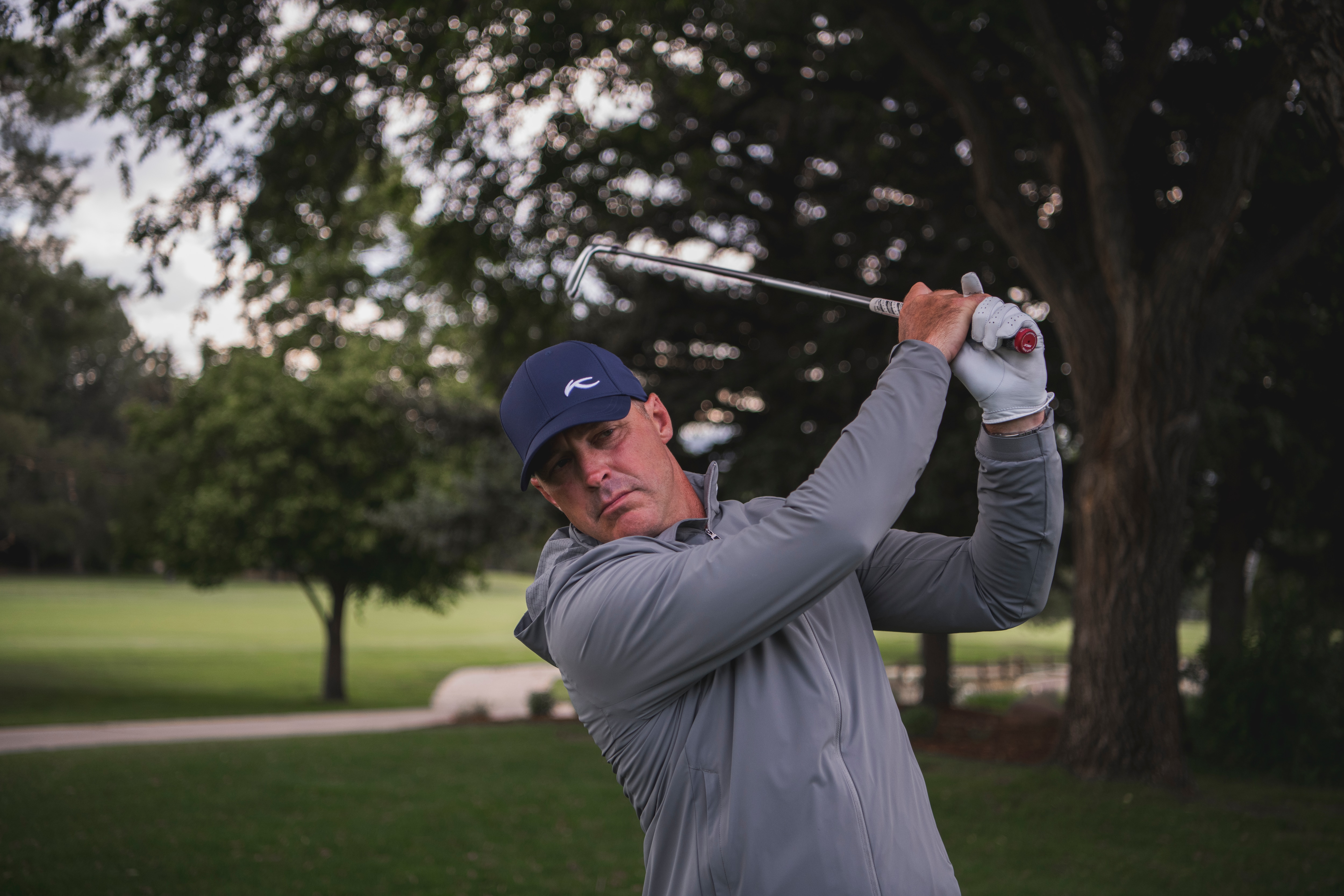 A man wearing a grey KJUS jacket and blue KJUS cap swinging a golf club.