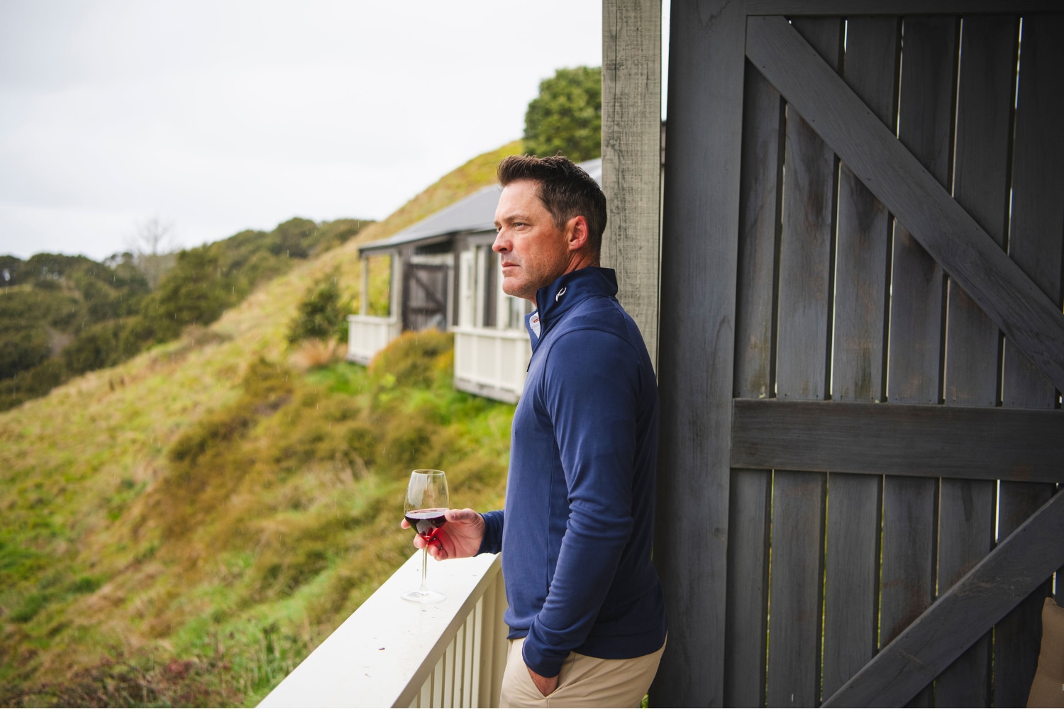A man wearing the KJUS Kallen Half-Zip in blue standing on a wooden proch holding a glass of red wine.